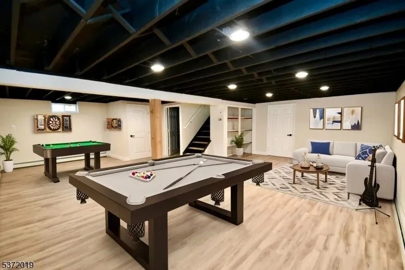 Basement recreation room with pool table, dartboard, with seating area including a white sofa, coffee table, and wall art, illuminated by ceiling lights.