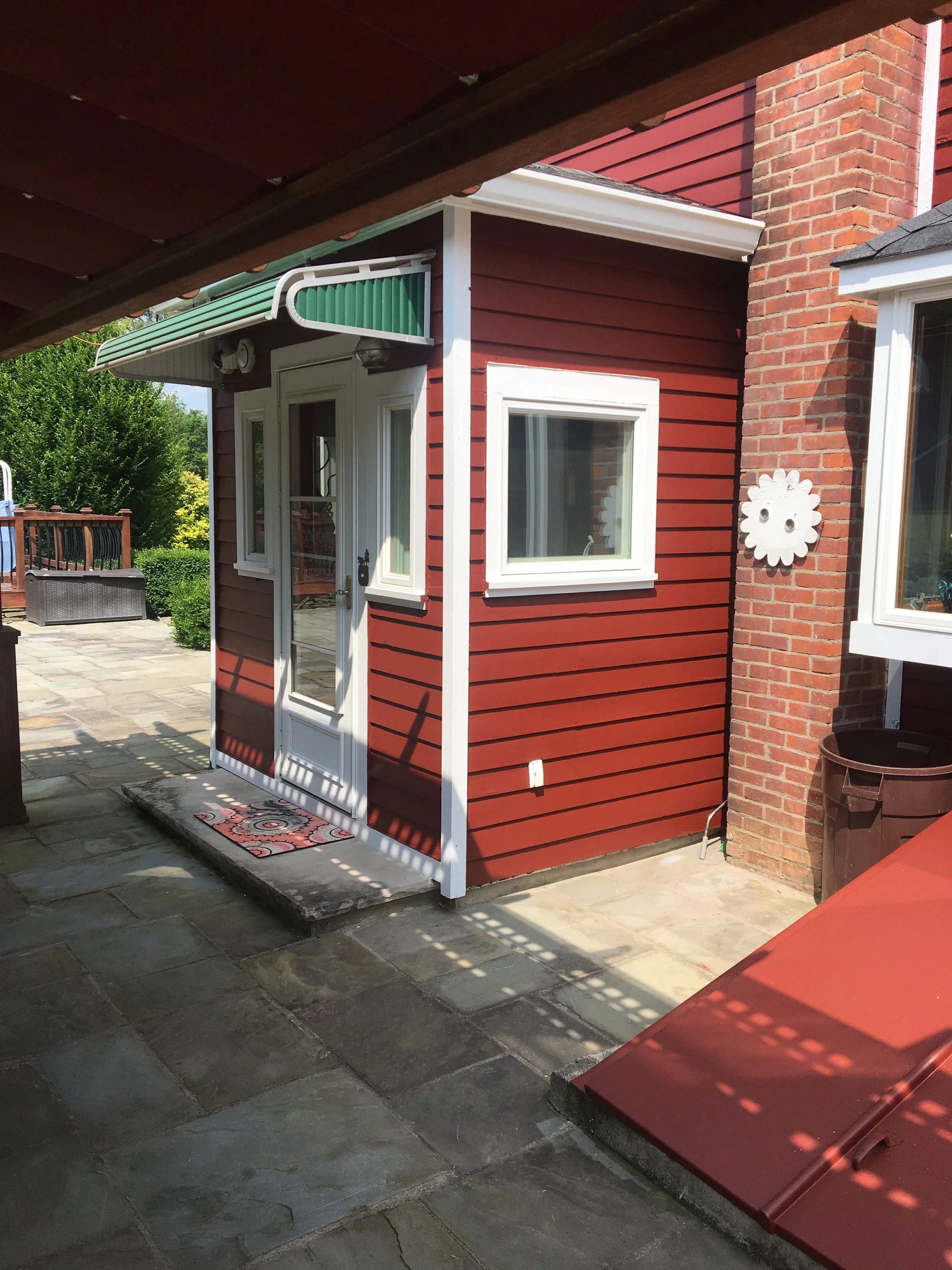 View of a red wooden shed with a white door and window, situated on a stone patio with outdoor furniture and greenery in the background.