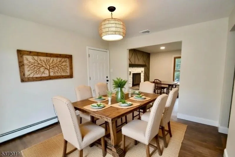 Dining room with six beige upholstered chairs around a wooden table set with plates, glasses, and green plants in a vase, hardwood floors, wall art, and a modern light fixture