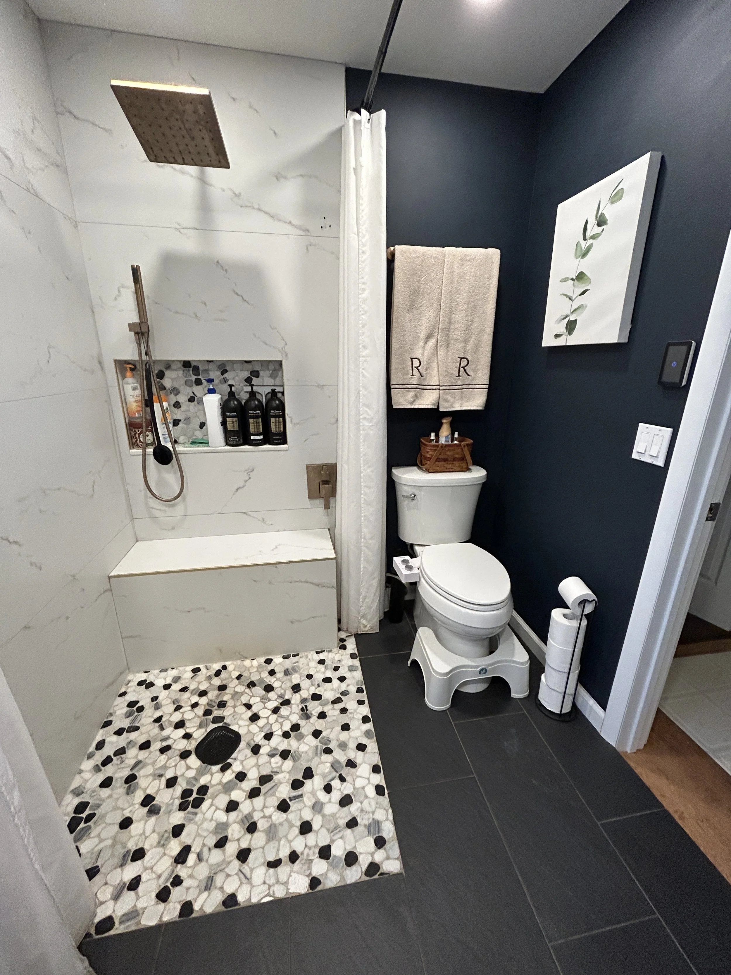 Bathroom with a walk-in shower featuring a gray and white tiled wall, a black and white pebble floor, a white toilet with a stool underneath, a dark navy wall with a framed leaf print, a towel rack with two beige towels, and a small wooden basket on 