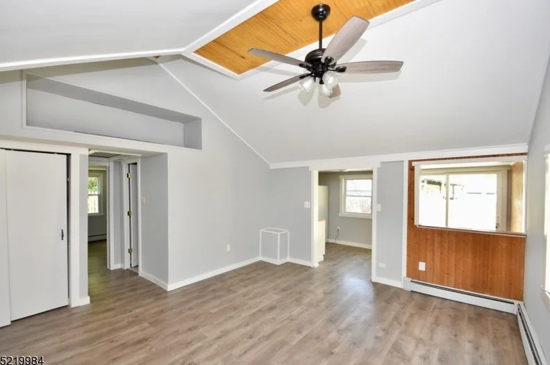 Empty living room with white walls, wood ceiling accents, a ceiling fan, hardwood flooring, and large windows.