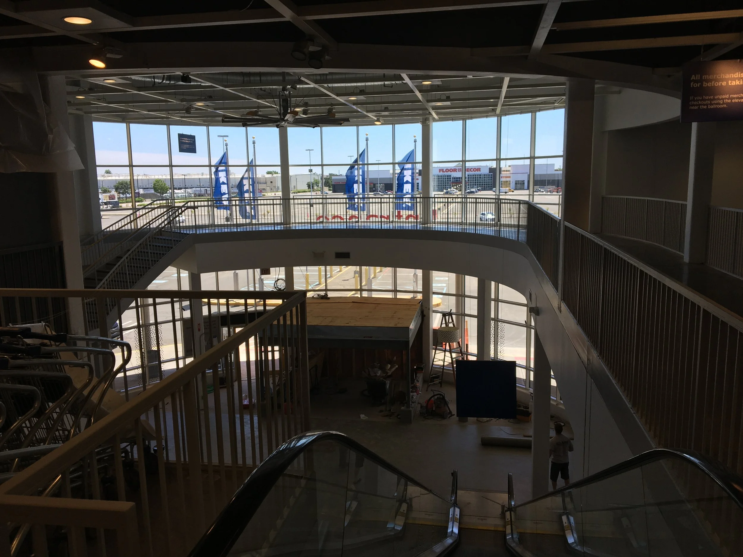 Interior view of a multi-level retail store with escalators, railing, large windows, and flags outside. Some signs and parked cars are visible through the windows.