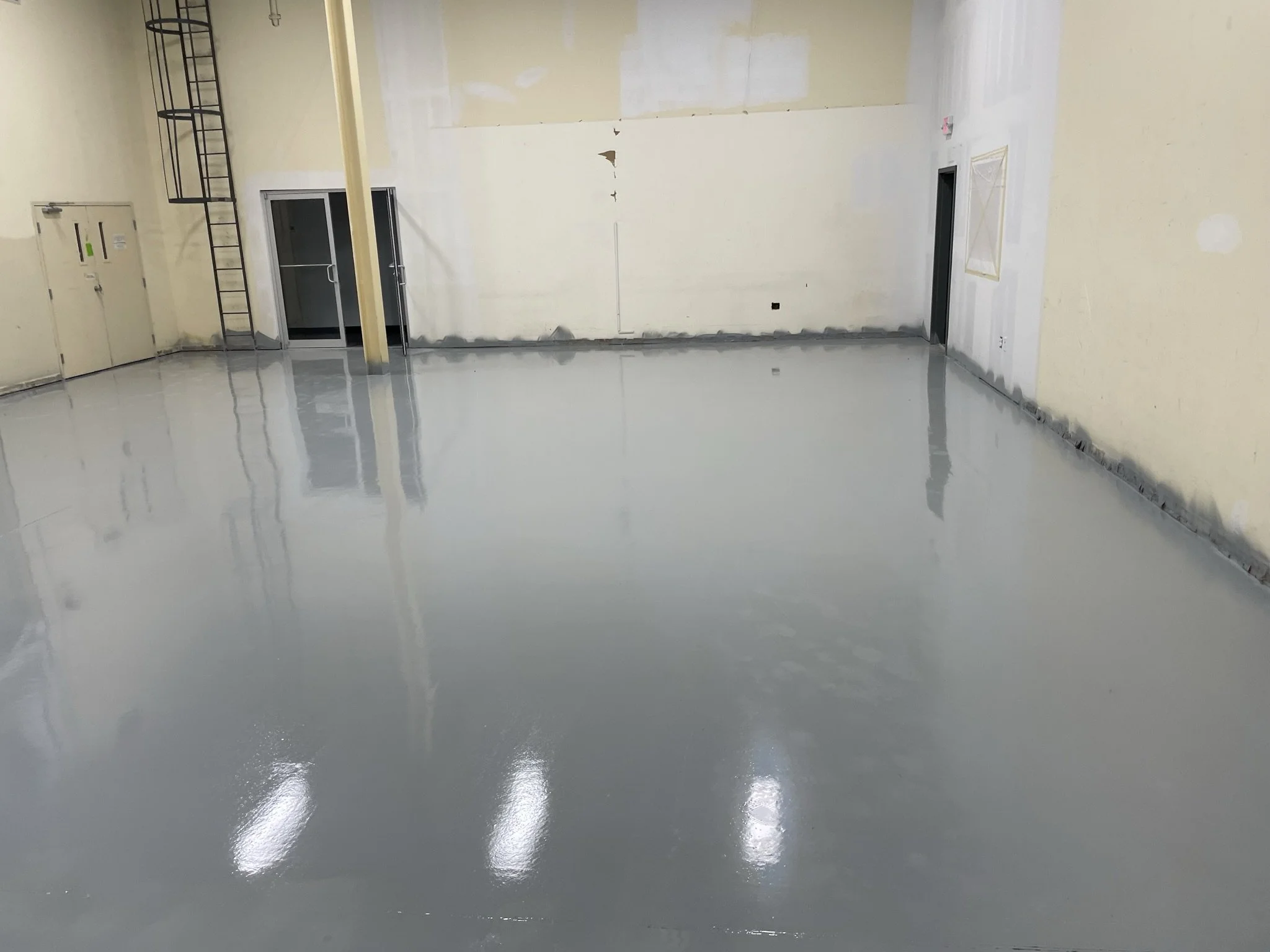 Empty indoor space with freshly painted shiny light gray floor, beige walls, and a few doors and ladders visible.