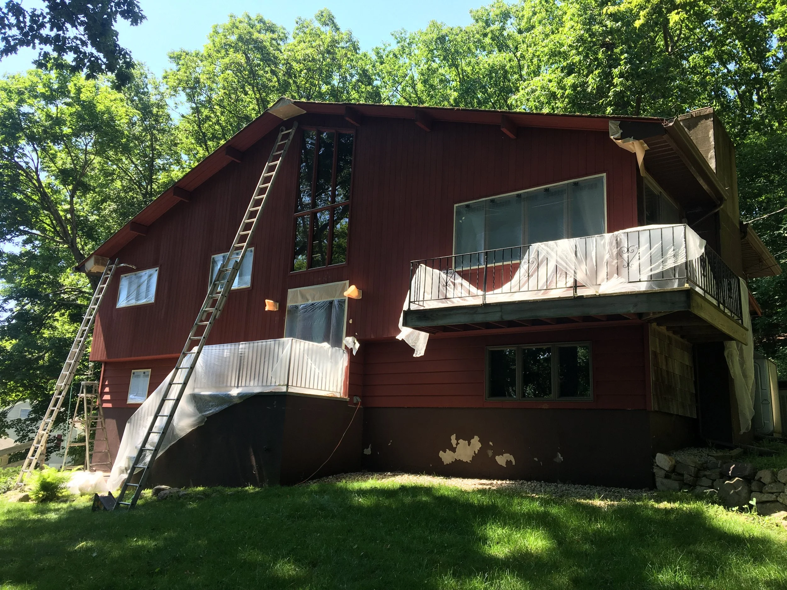 A two-story house with a red exterior is undergoing exterior renovation, with white protective sheeting draped over the balcony and windows, and ladders positioned against the house. The house is surrounded by green trees and grass.