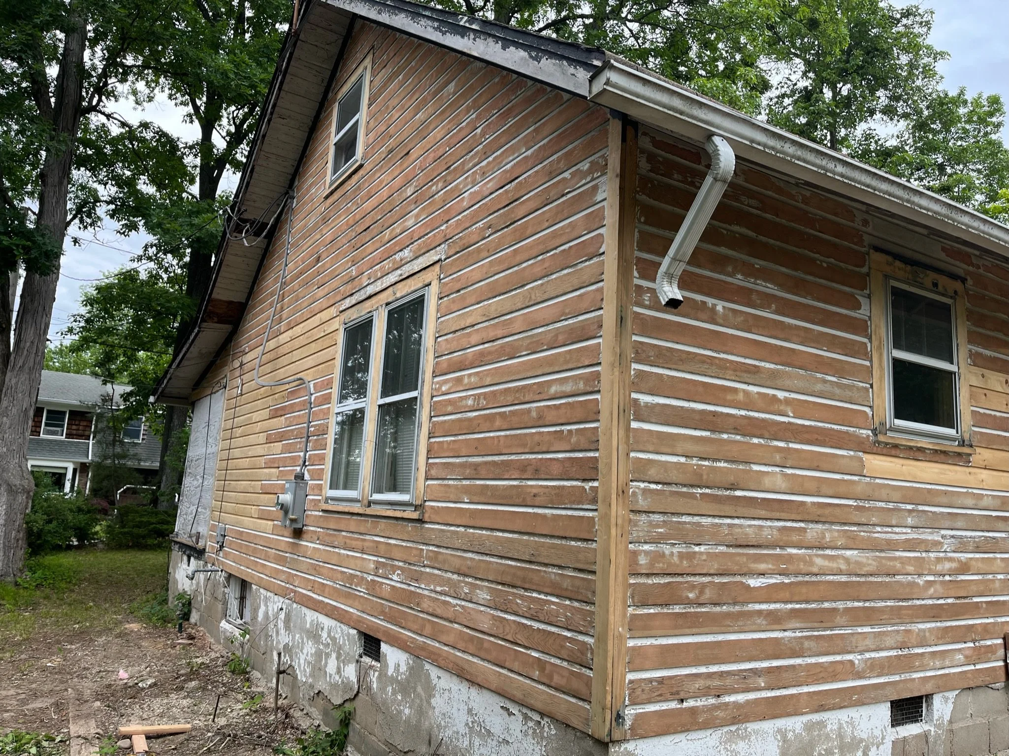 Back view of a house undergoing siding renovation, with exposed wood siding and a partially replaced section, surrounded by trees.