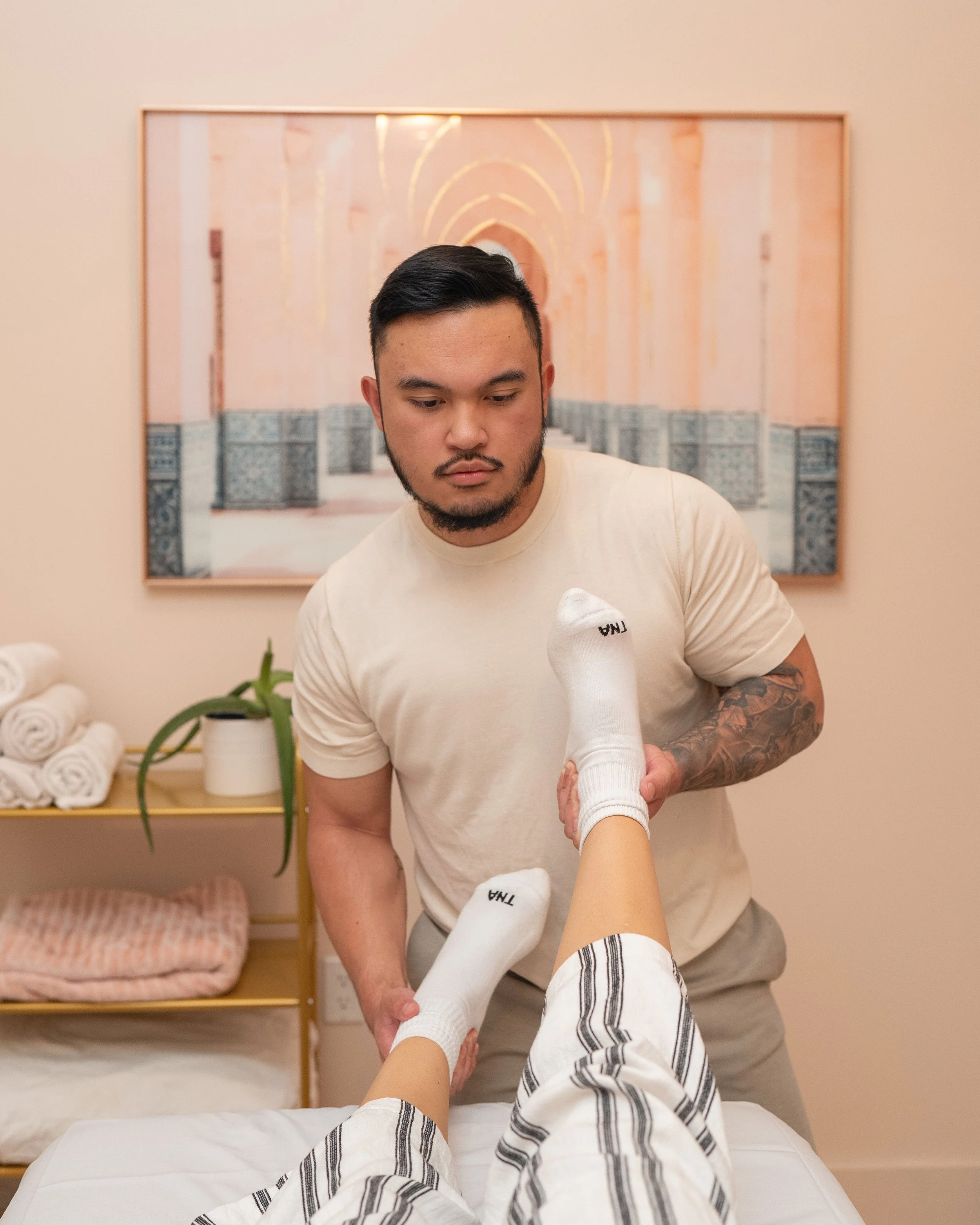 A man is giving a leg massage or treatment to a person lying on a massage table, with the person's legs elevated. The setting appears to be a spa or therapy room, with rolled towels and a plant on a nearby shelf, and framed artwork on the wall.