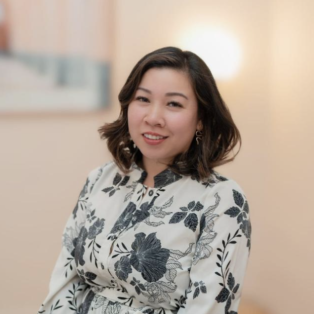 A woman with shoulder-length dark hair smiling, wearing a white blouse with black floral print, in a softly lit indoor setting.