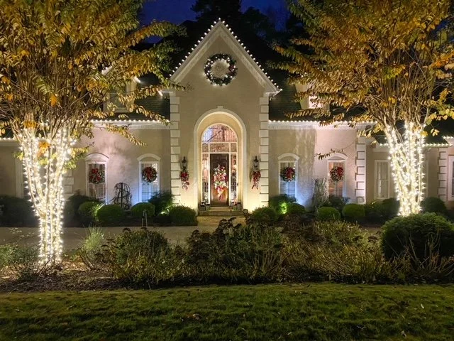 House decorated with Christmas wreaths and Christmas lights on trees in front yard at night.