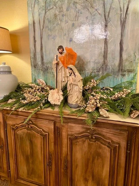 Religious figurines of Jesus and Mary on a wooden cabinet, surrounded by floral decorations, with a lake and trees painted on a wall in the background.