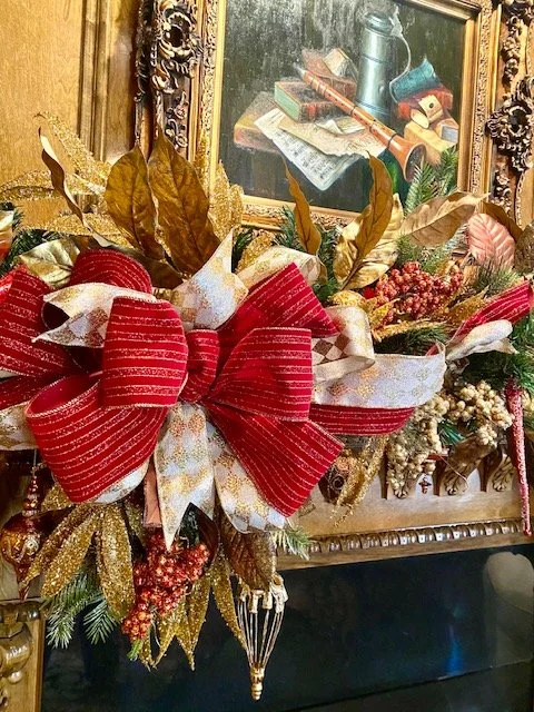 Christmas floral arrangement with large red and gold bows, gold and brown leaves, and festive greenery, placed in front of a framed painting on a wooden surface.