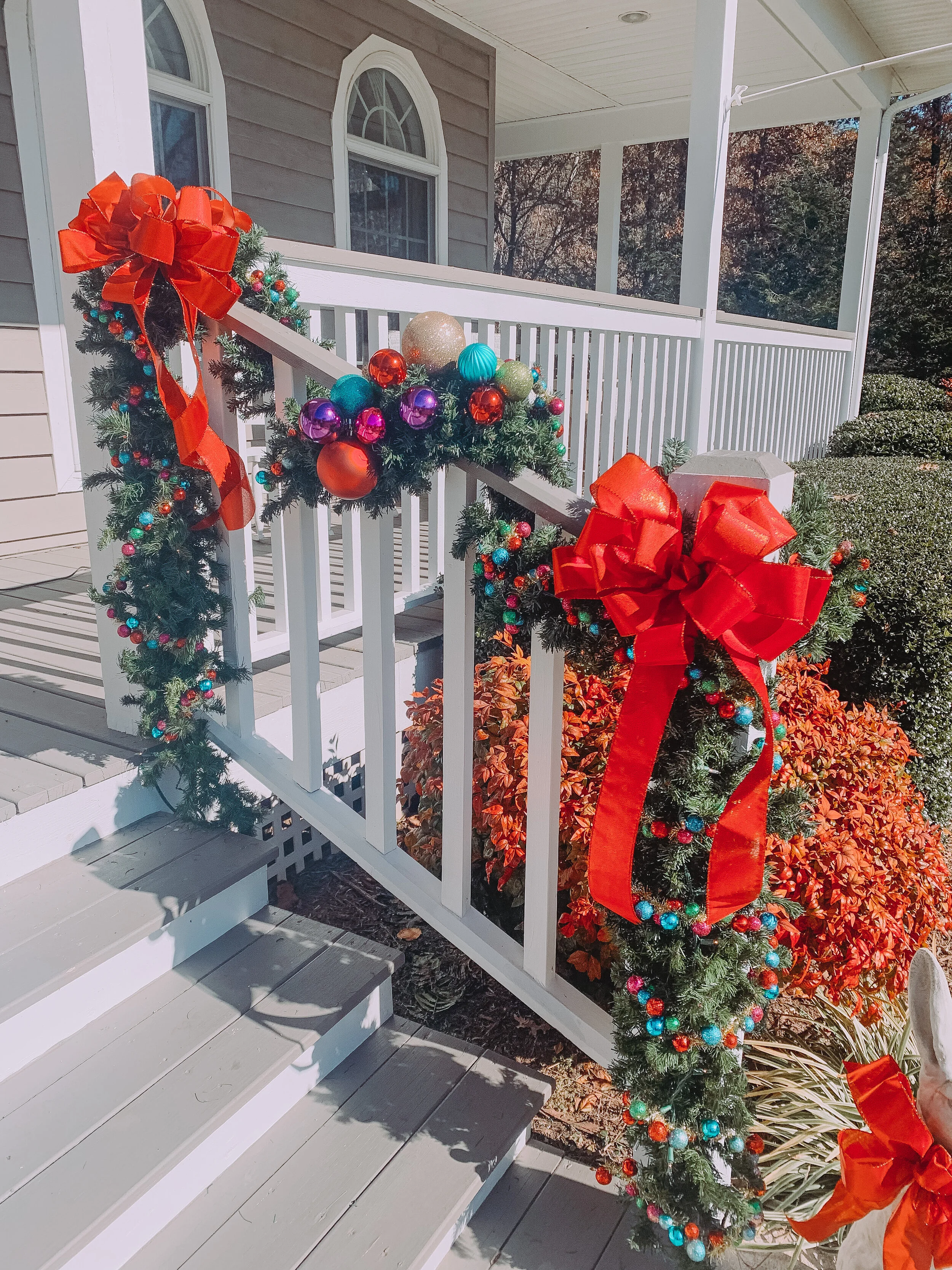Decorated front porch railing with Christmas garlands, large red bows, colorful ornaments, and a house with gray siding and white trim.
