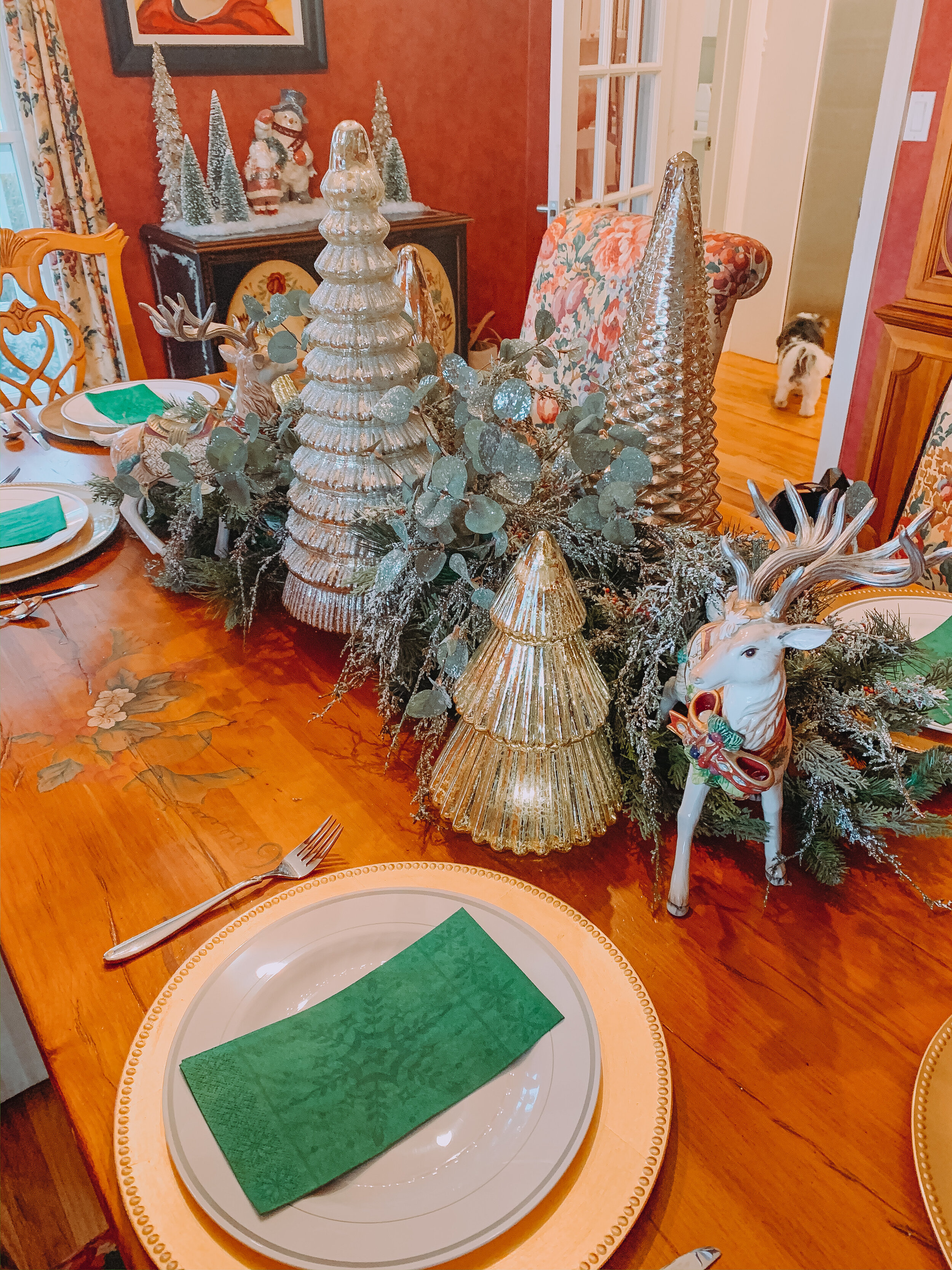 Decorative Christmas centerpiece on a dining table, featuring glass Christmas tree and reindeer figurines, silver and gold ornaments, and green foliage, with place settings and green napkins.