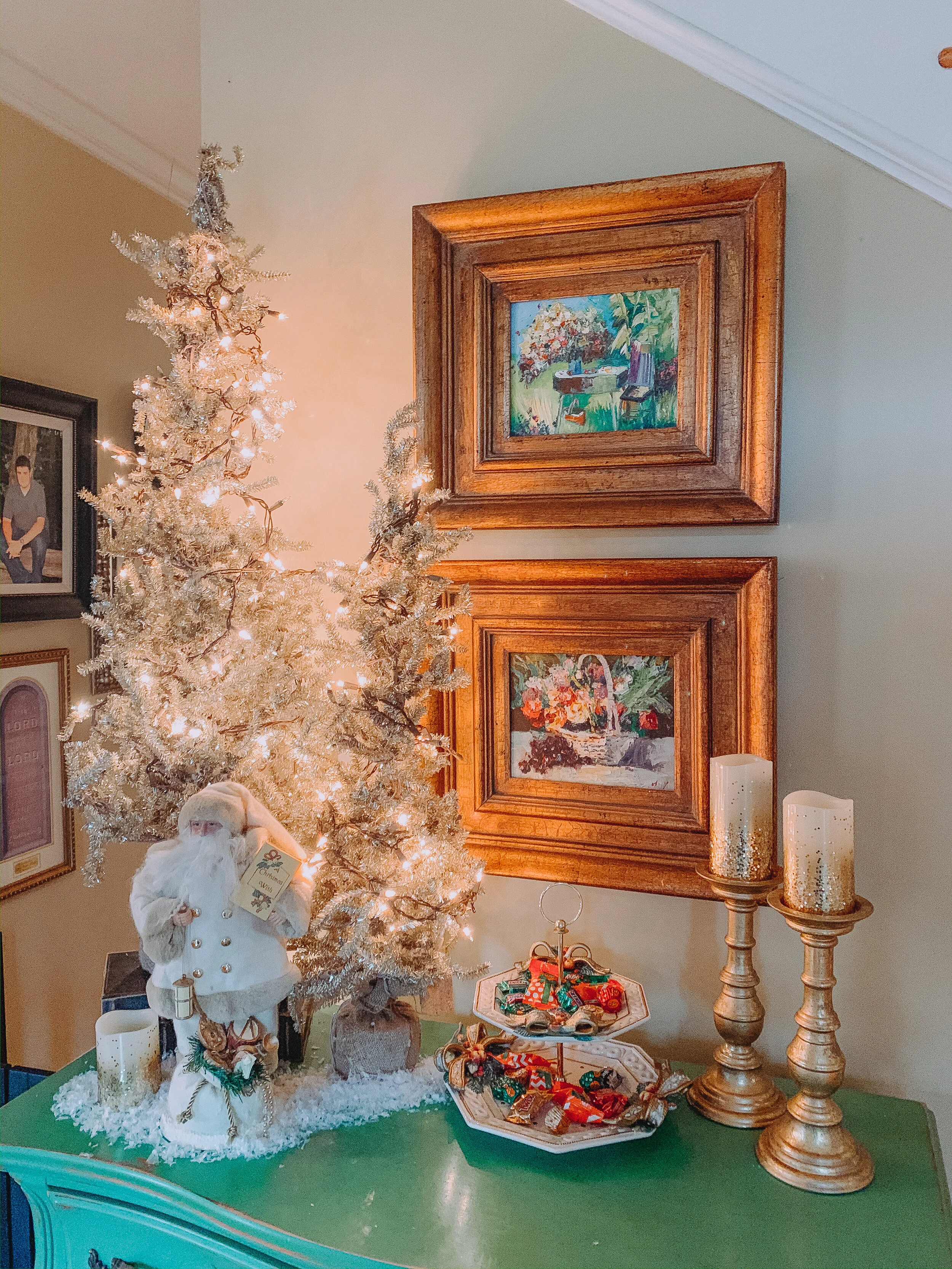Decorative Christmas scene with a white flocked Christmas tree adorned with warm white lights, a Santa Claus figurine holding a gift, two gold candle holders with white candles, a tiered tray with small wrapped gifts, and framed paintings hanging on 