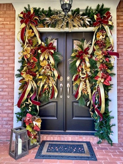 Decorative Christmas wreaths and ribbons on a black front door, with holiday greenery and bows on each side, and a lantern with a candle on the brick porch.