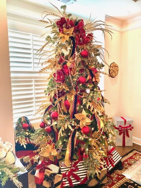 Decorated Christmas tree with red ornaments, gold ribbons, and gold leaves, surrounded by wrapped presents in a living room with white shutters and a wall clock.