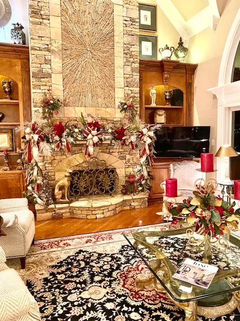 Living room with fireplace decorated for Christmas, adorned with red and gold garlands, candles, and flowers. There is a television, a glass coffee table with magazines, and armchairs.