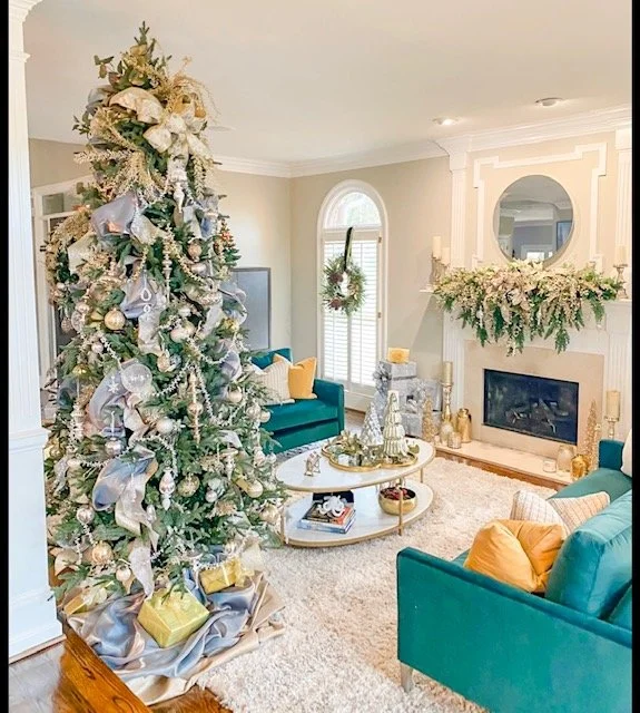 Decorated Christmas tree with gold and silver ornaments and ribbons in a cozy living room with teal sofas, a fireplace decorated with greenery, and holiday decorations.