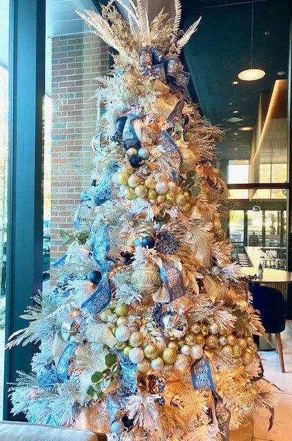 Decorated Christmas tree with gold, blue, and white ornaments and ribbons inside a modern indoor space.