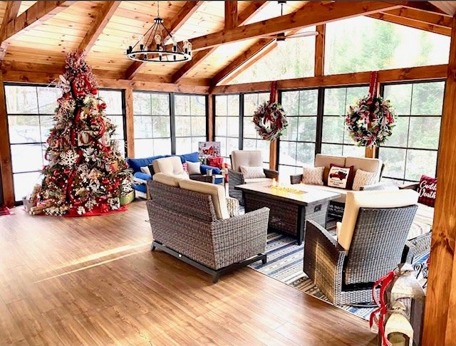 A sunlit indoor living room decorated for Christmas with a large decorated Christmas tree, wreaths hanging on the windows, and holiday pillows on the furniture.
