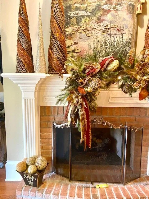 Decorative autumn arrangement on a brick fireplace mantel with twined pillars and floral artwork above, gold-colored orbs in a black container on the floor in front.