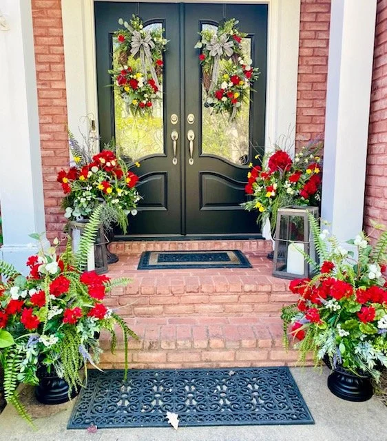Decorated front porch with black double doors adorned with floral wreaths, and large floral arrangements with red, white, yellow flowers, and green foliage on either side.