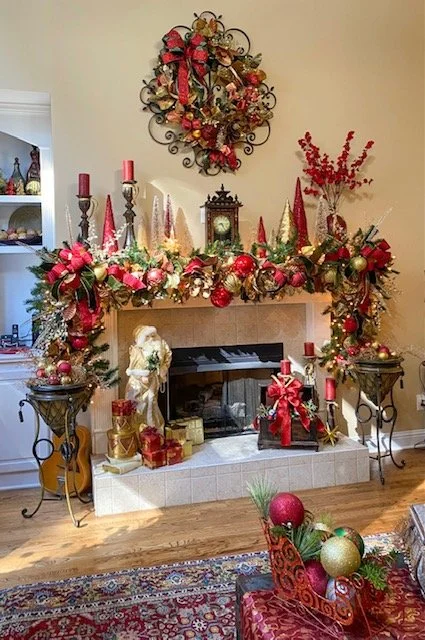 Festive Christmas fireplace decorated with red and gold ornaments, ribbons, candles, and floral arrangements, with stockings hanging and holiday decorations on nearby tables.
