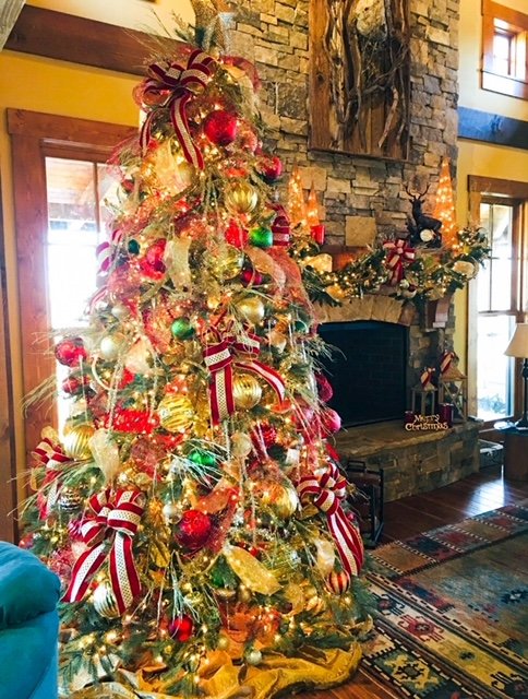Christmas tree decorated with gold, red, and green ornaments, ribbons, and lights in a cozy living room with stone fireplace and mantel decorated with ornaments and greenery.