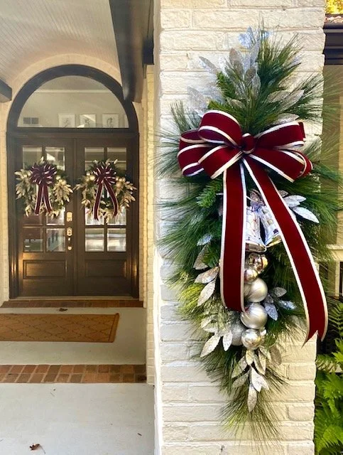 Decorative Christmas wreath with silver ornaments, large red and white bow, evergreen branches, and silver leaves on a brick wall near an entrance door with additional wreaths.
