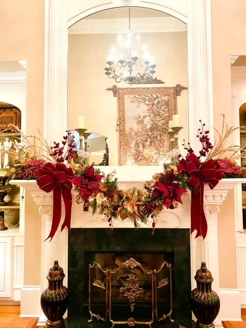 Festive decorated fireplace mantel with red bows, greenery, candles, and a tapestry reflected in the mirror above.