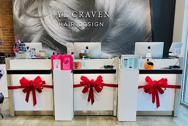 Salon reception desk decorated with large red bows and ribbons, with computer monitors and hair products visible on the counters, and a black and white hair-themed wall sign reading 'JYL CRAVEN HAIR DESIGN'.