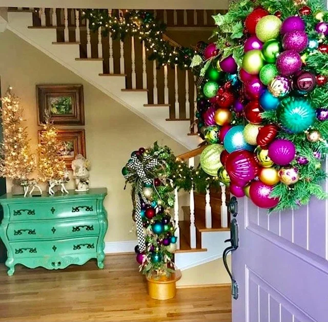 Festively decorated staircase landing with a large colorful Christmas ornament wreath near the door, a green dresser with gold accents, small Christmas trees, and reindeer figurines, all illuminated with holiday lights.