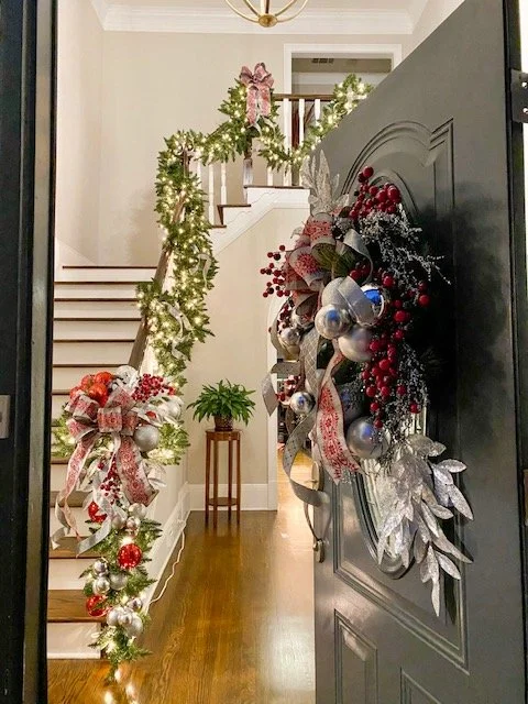 Holiday decorated front door with silver ornaments, red berries, glittery leaves, ribbons, and a green garland with ornaments and ribbons on a staircase railing inside a home.