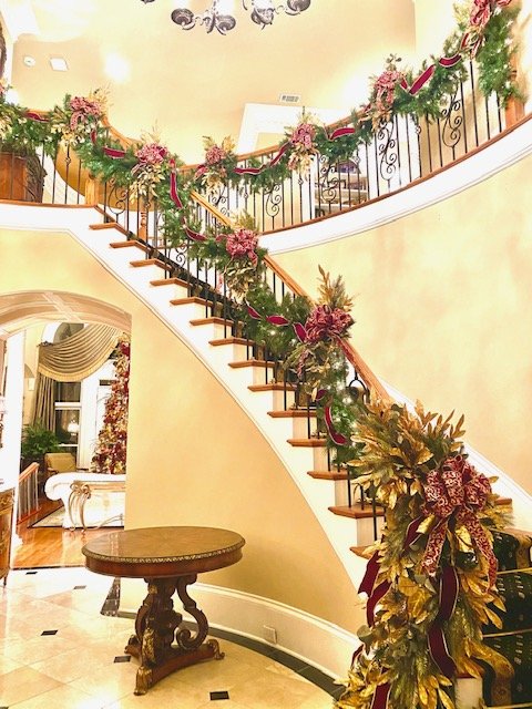 Indoor staircase decorated with Christmas garlands and flowers for the holiday season, featuring a curved wooden handrail and ornate newel post, with a seating area and window in the background.