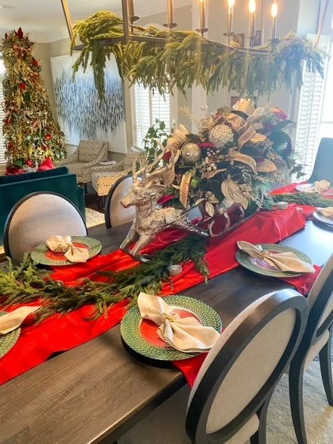 Christmas dining table decorated with a red table runner, gold and silver foliage, reindeer figurines, candles, and place settings with cloth napkins and green chargers.