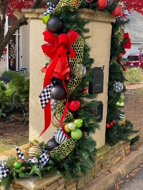 Decorated outdoor mailbox with Christmas ornaments, red bow, and festive ribbons surrounded by greenery.