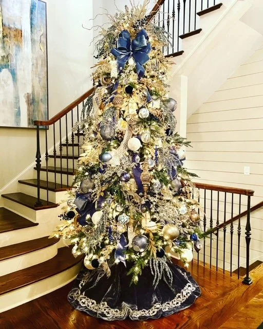 Christmas tree decorated with silver, blue, and gold ornaments, ribbons, and lights, placed in front of staircase with wooden railing.
