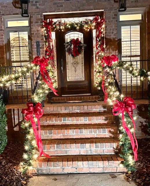 Front porch decorated for Christmas with garland, red ribbons, lights, and a wreath on the door.