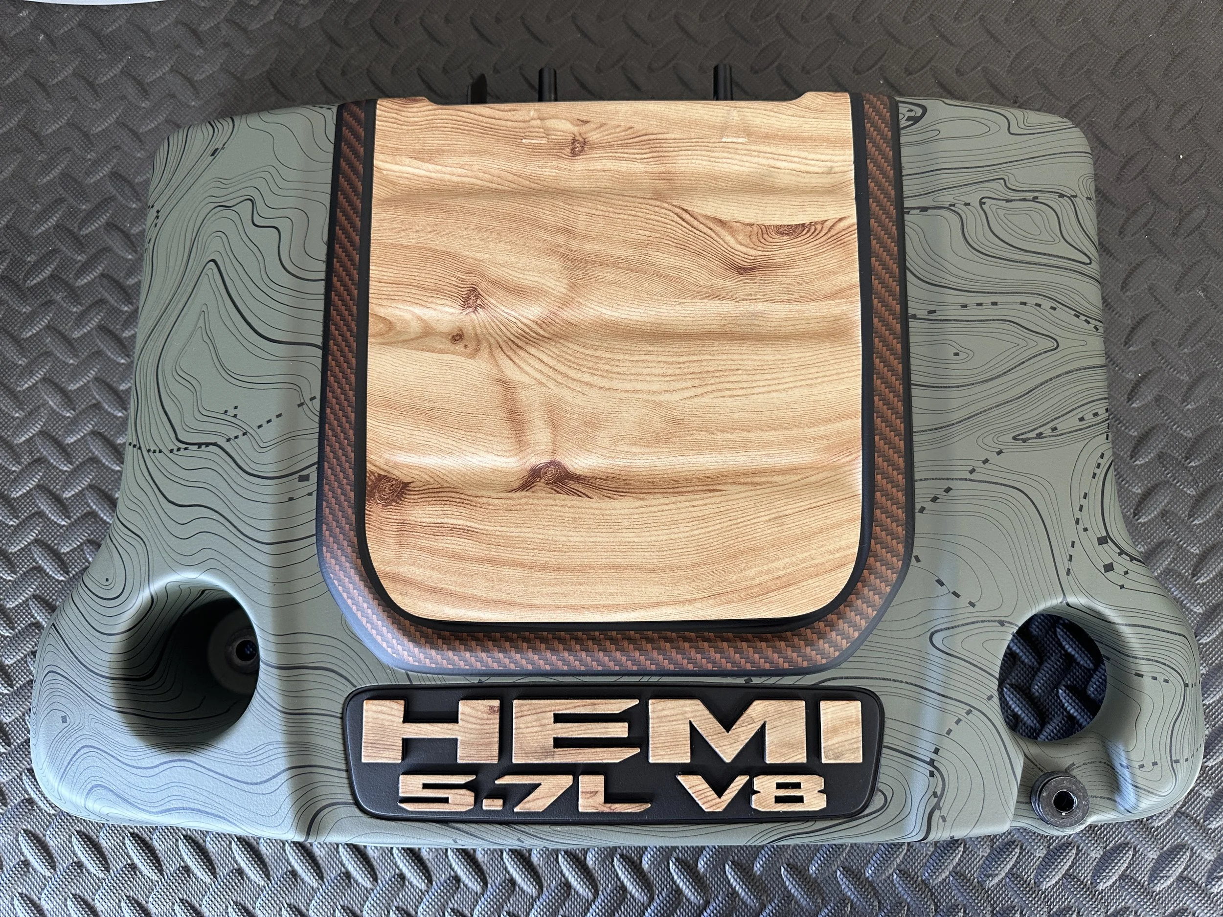 A close-up view of a camouflaged engine cover with a wooden accent and a 5.7L V8 HEMI badge, placed on a textured metallic surface.