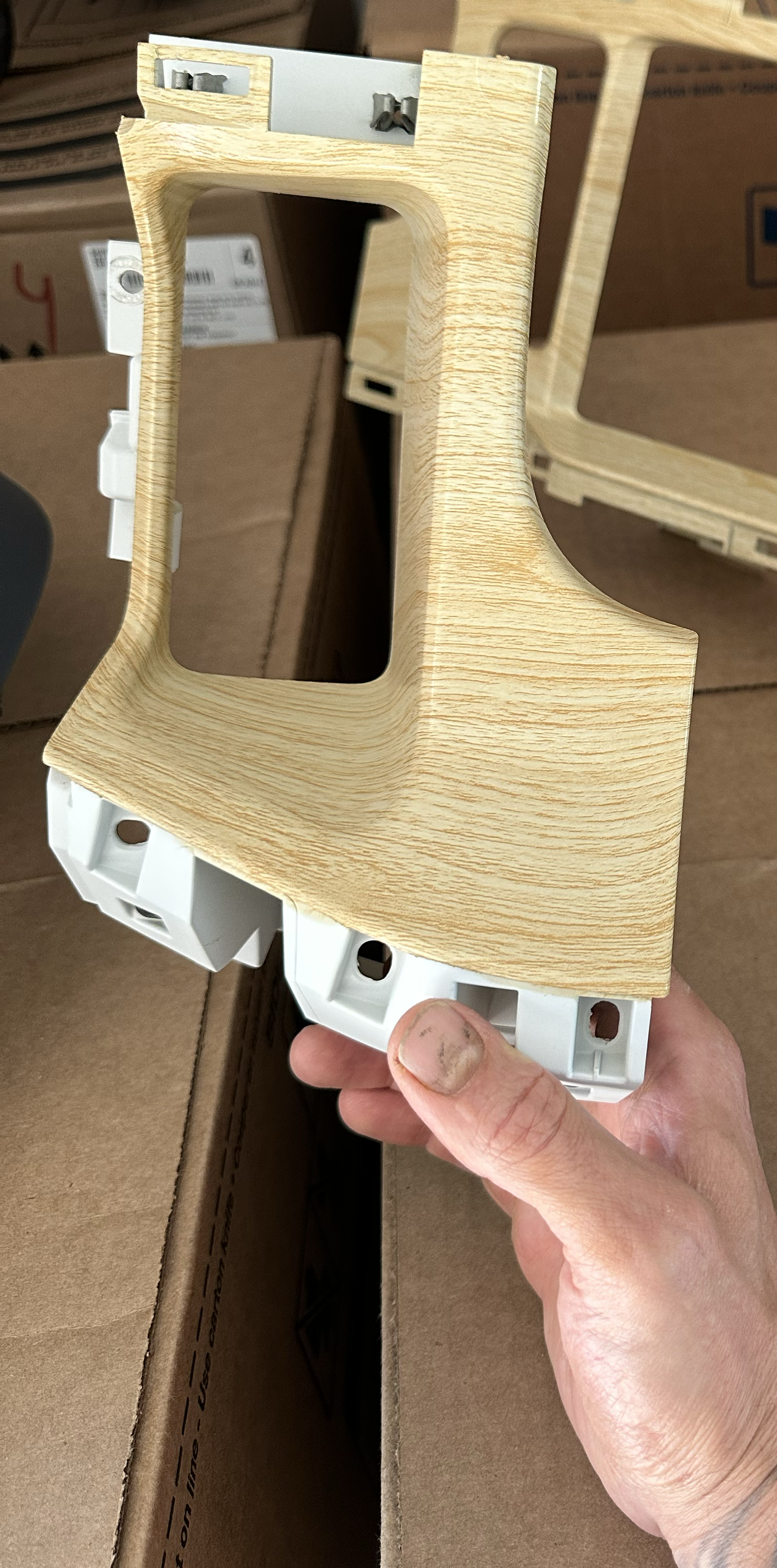 Person holding a wooden and plastic frame, possibly a car dashboard component, with a large opening in the center and mounting points around the edges, surrounded by cardboard boxes.