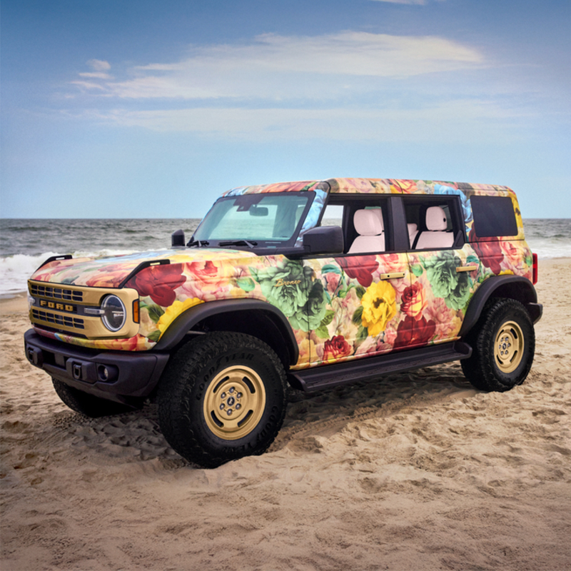 A Ford SUV with a colorful floral wrap parked on a sandy beach near the ocean under a partly cloudy sky.