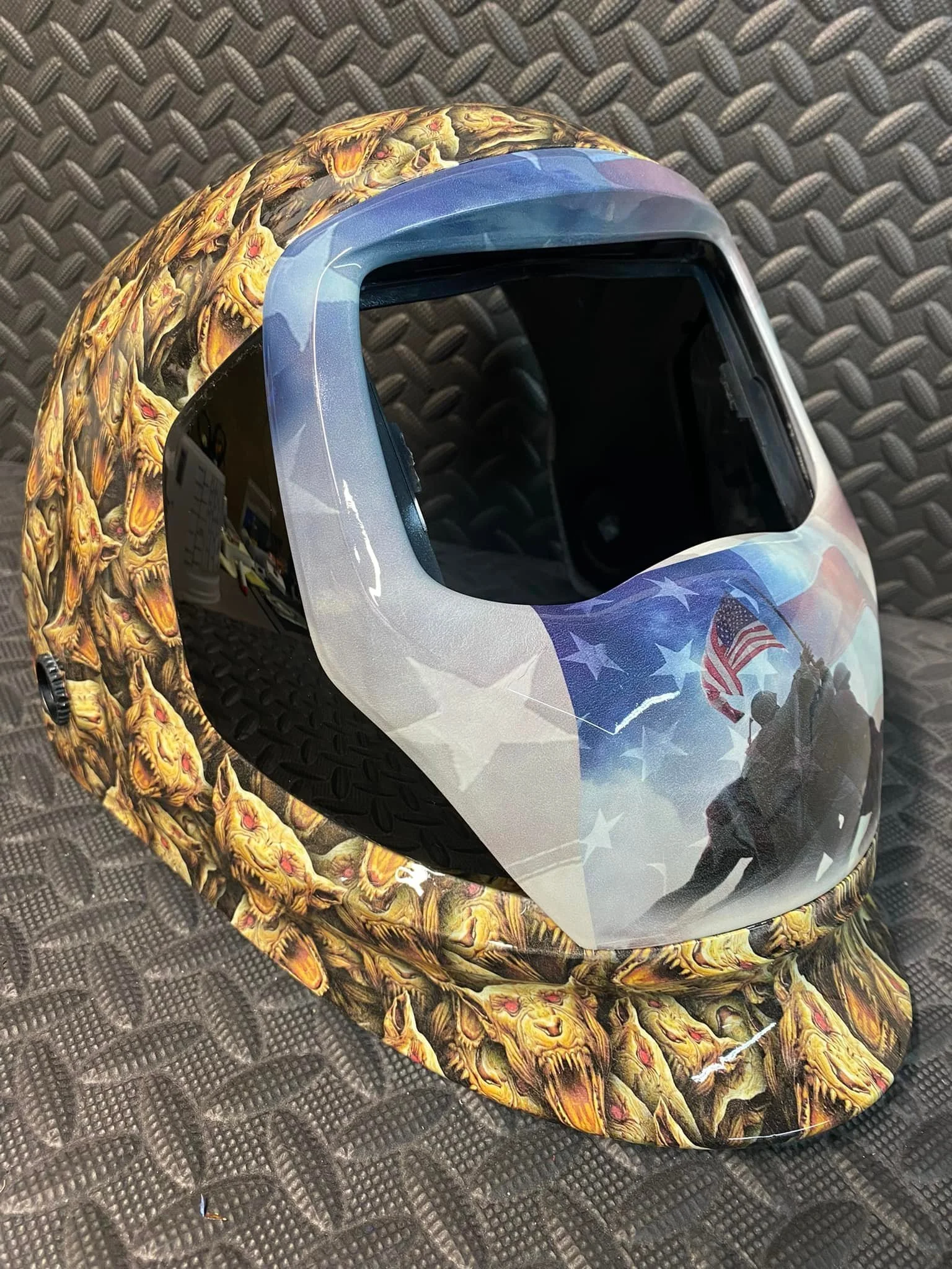 A welding helmet with an American flag design, featuring a quote of a soldier holding an American flag, resting on a snake print hat on a black textured surface.