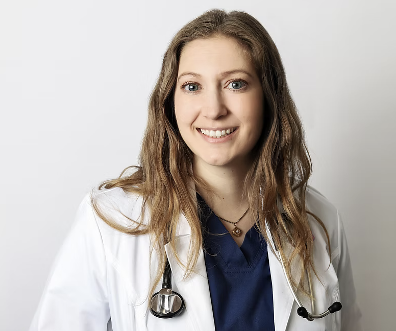 A young female doctor smiling, wearing a white coat and navy scrubs, with a stethoscope around her neck, standing against a plain light background.