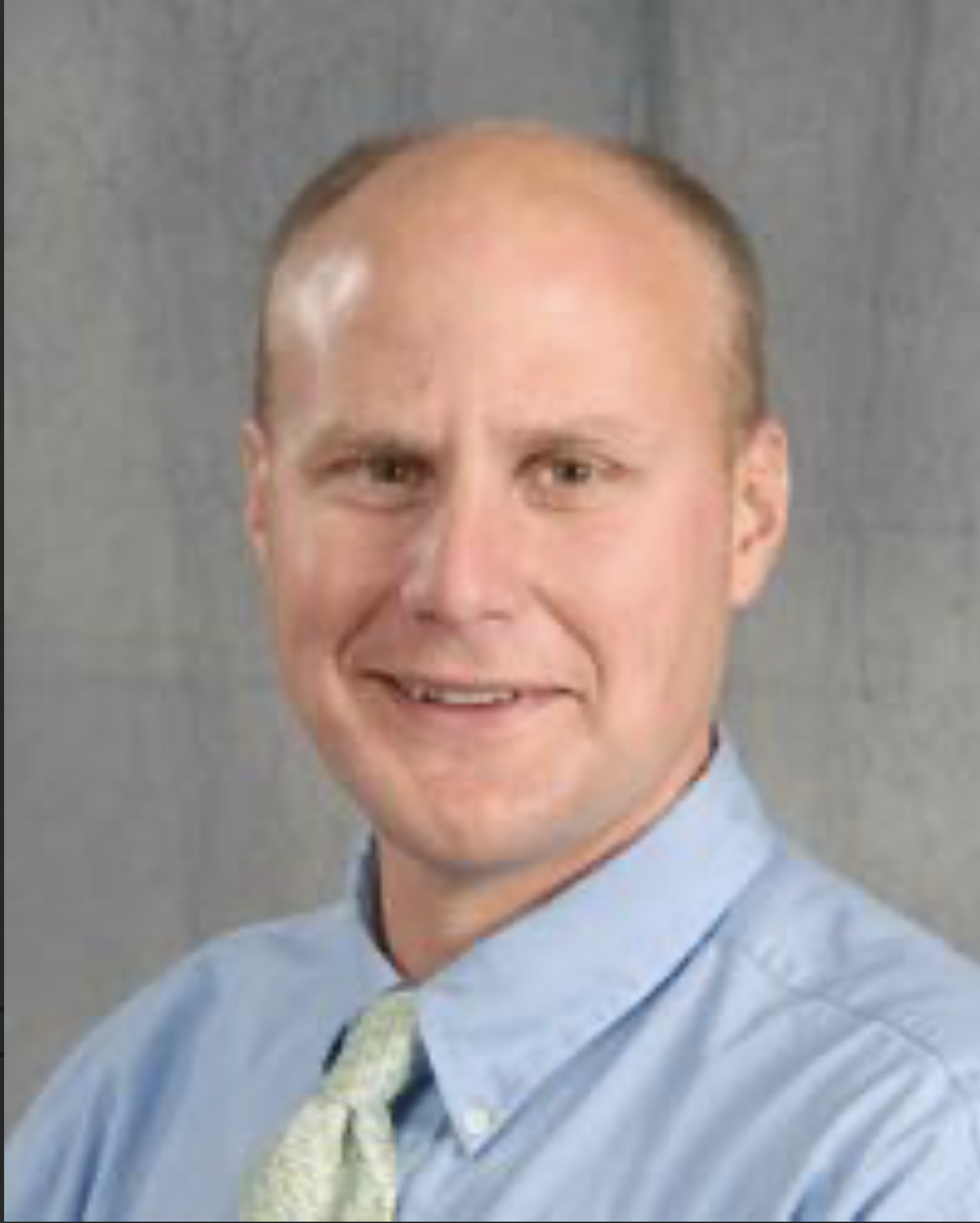 Headshot of a smiling man in a blue dress shirt and light-colored tie, against a gray background.