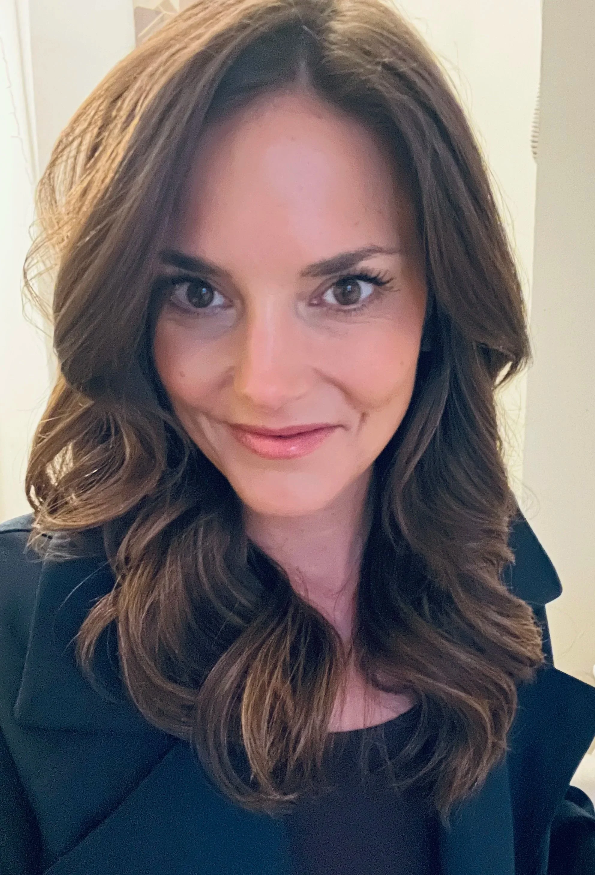 Woman with long brown hair wearing a black floral blouse, smiling, against a dark blue background.