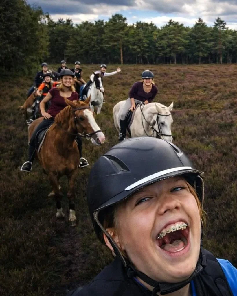 Selfie van een groep ruiters die rijden over een ruiterpad dwars door een heideveld met op de achtergrond een bosrand.