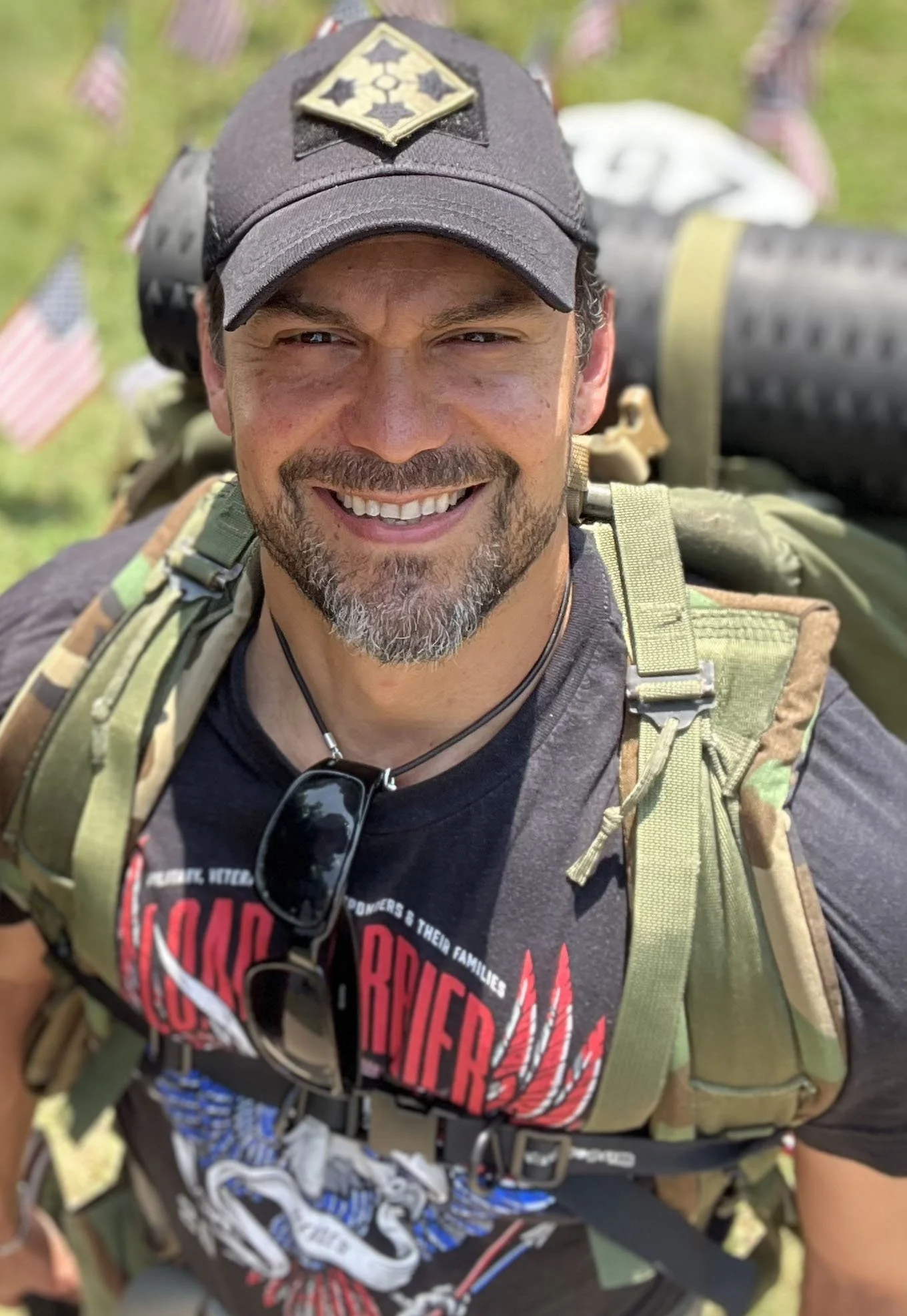 A smiling U.S. Army Veteran man wearing a black baseball cap with a graphic patch, sunglasses hanging from his shirt, a camouflage rucksack, and a black T-shirt with red and white graphics. He is outdoors with American flags in the background.