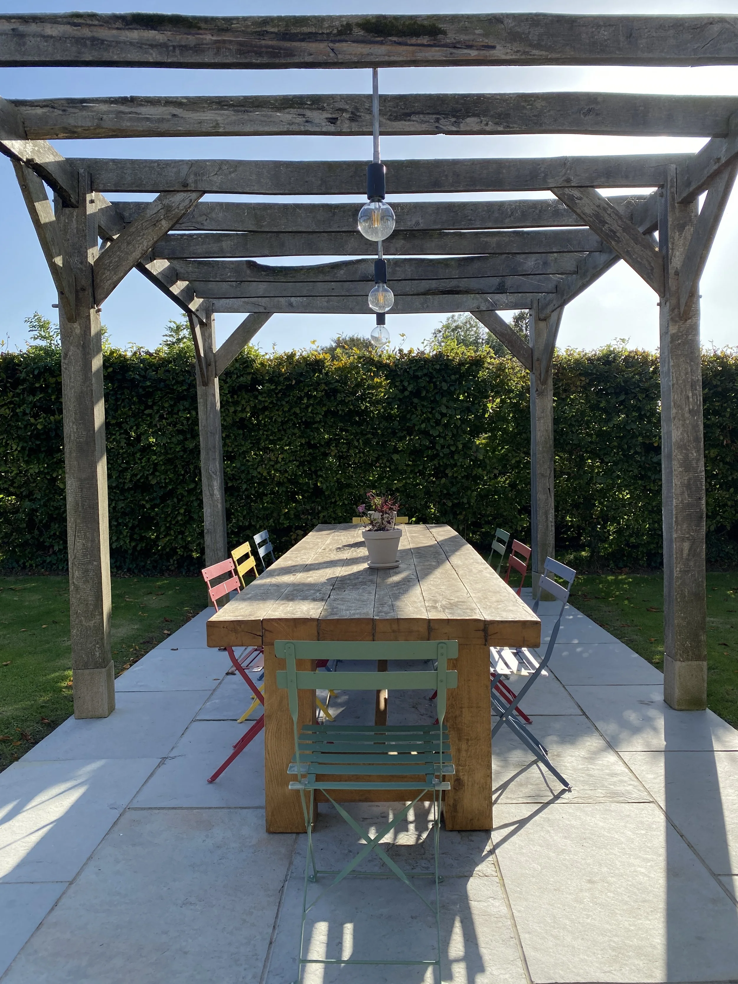 Outdoor dining area with a large wooden table, surrounded by colorful metal chairs, under a wooden pergola with hanging light bulbs, on a tiled patio with a background of a green hedge.