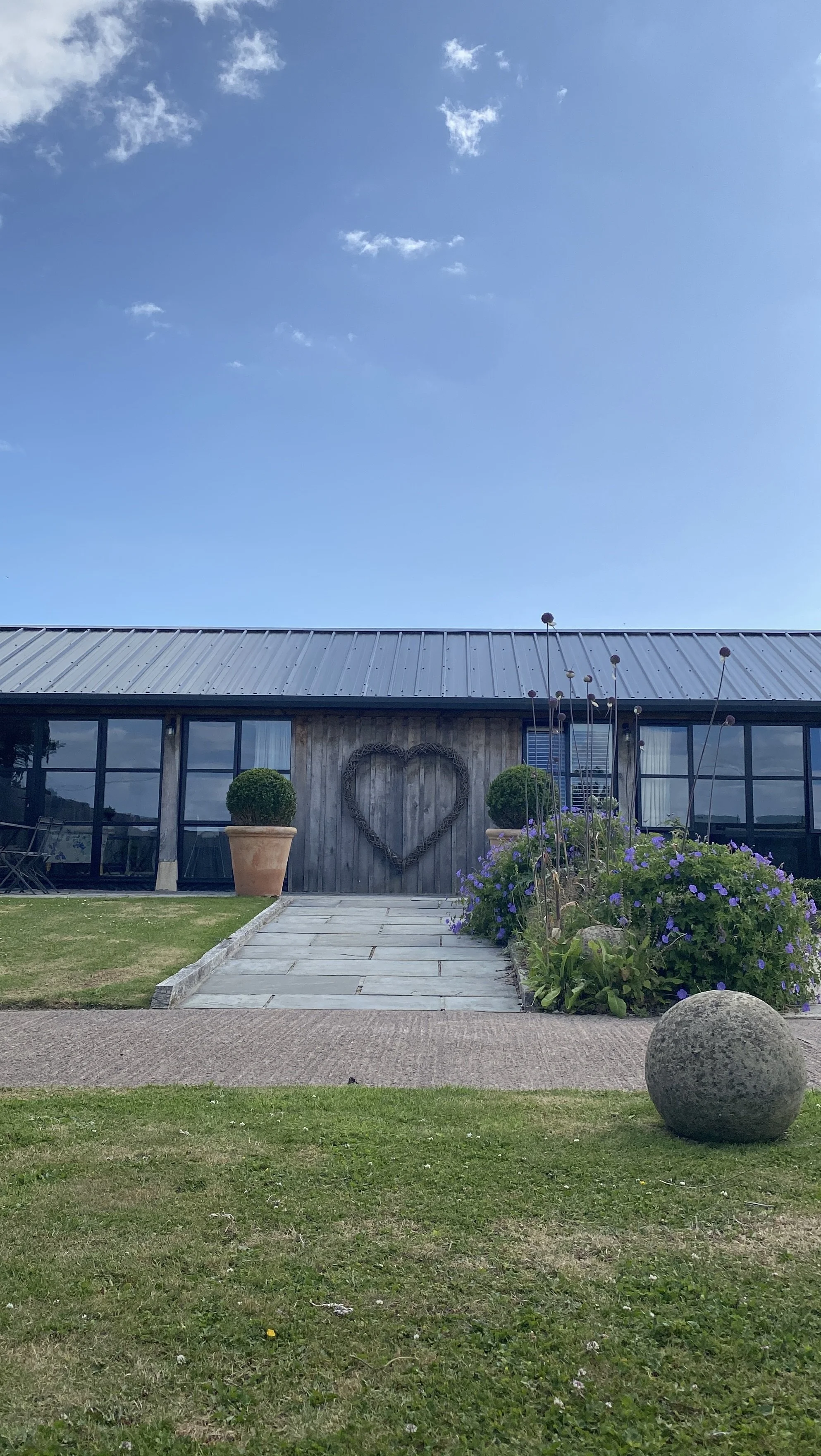 A building with a metal roof, glass windows, a heart-shaped decoration on the wall, potted plants, a flower bed with purple flowers, a large stone sphere, and a paved pathway leading to the entrance.