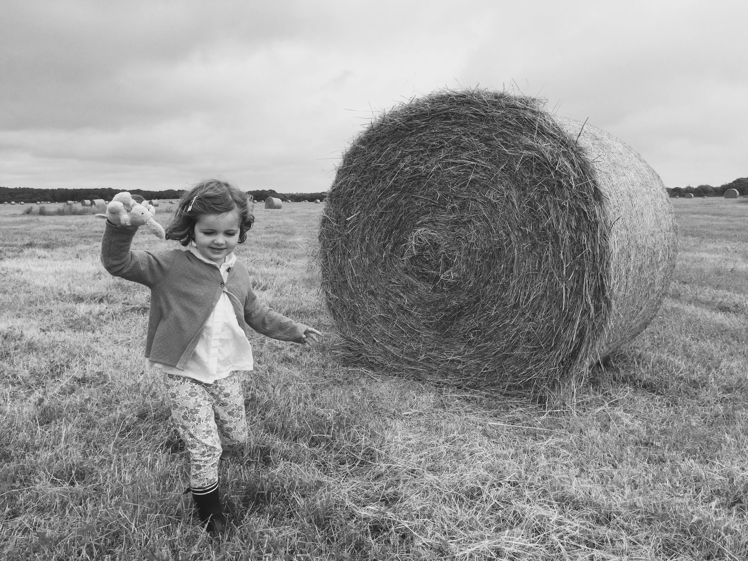 Black and white photo of a child playing near a hay bale in a field.