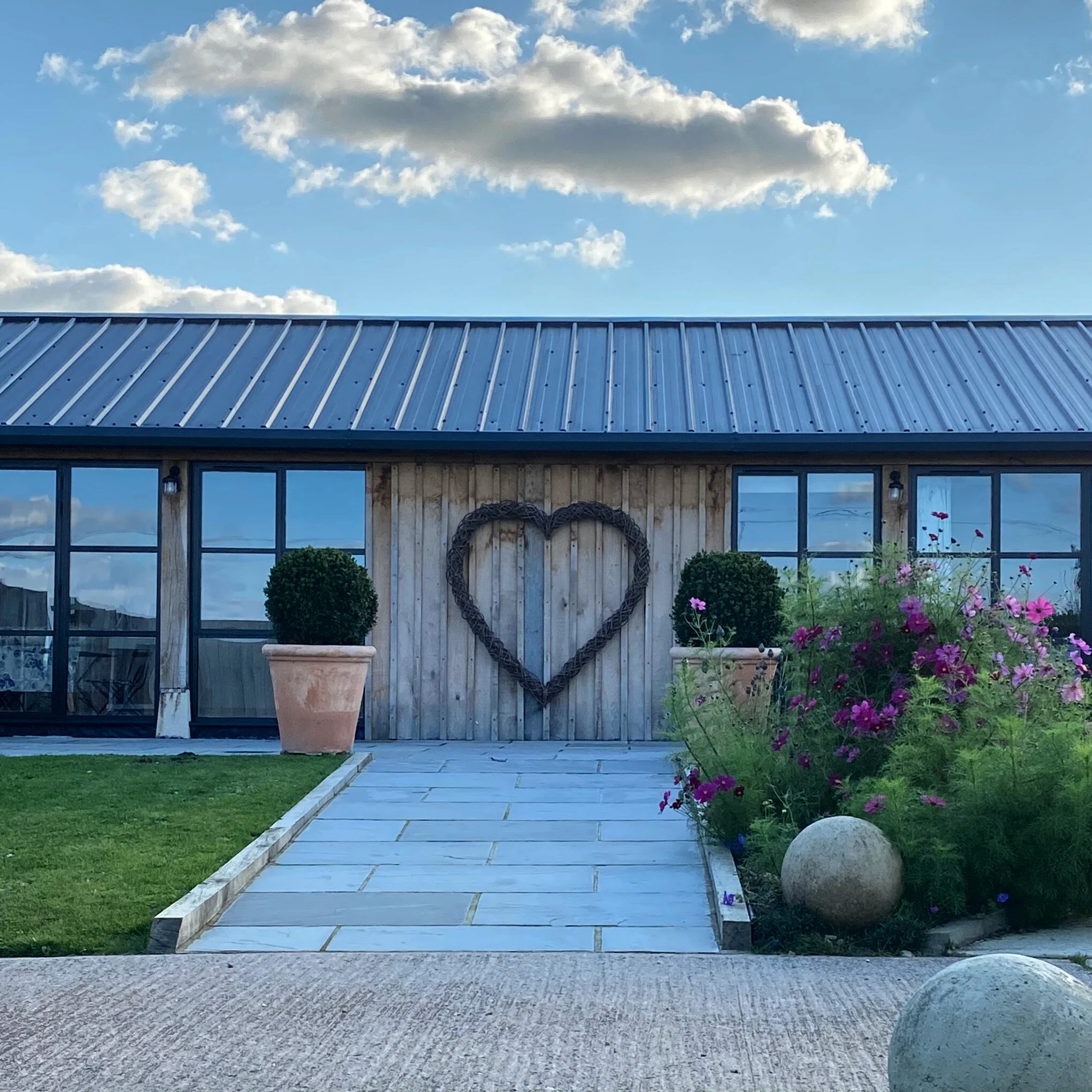 A building with glass windows and a wooden exterior, decorated with a large heart-shaped wreath on the wall, two potted plants on either side, flowering bushes, and a clear blue sky with some clouds overhead.